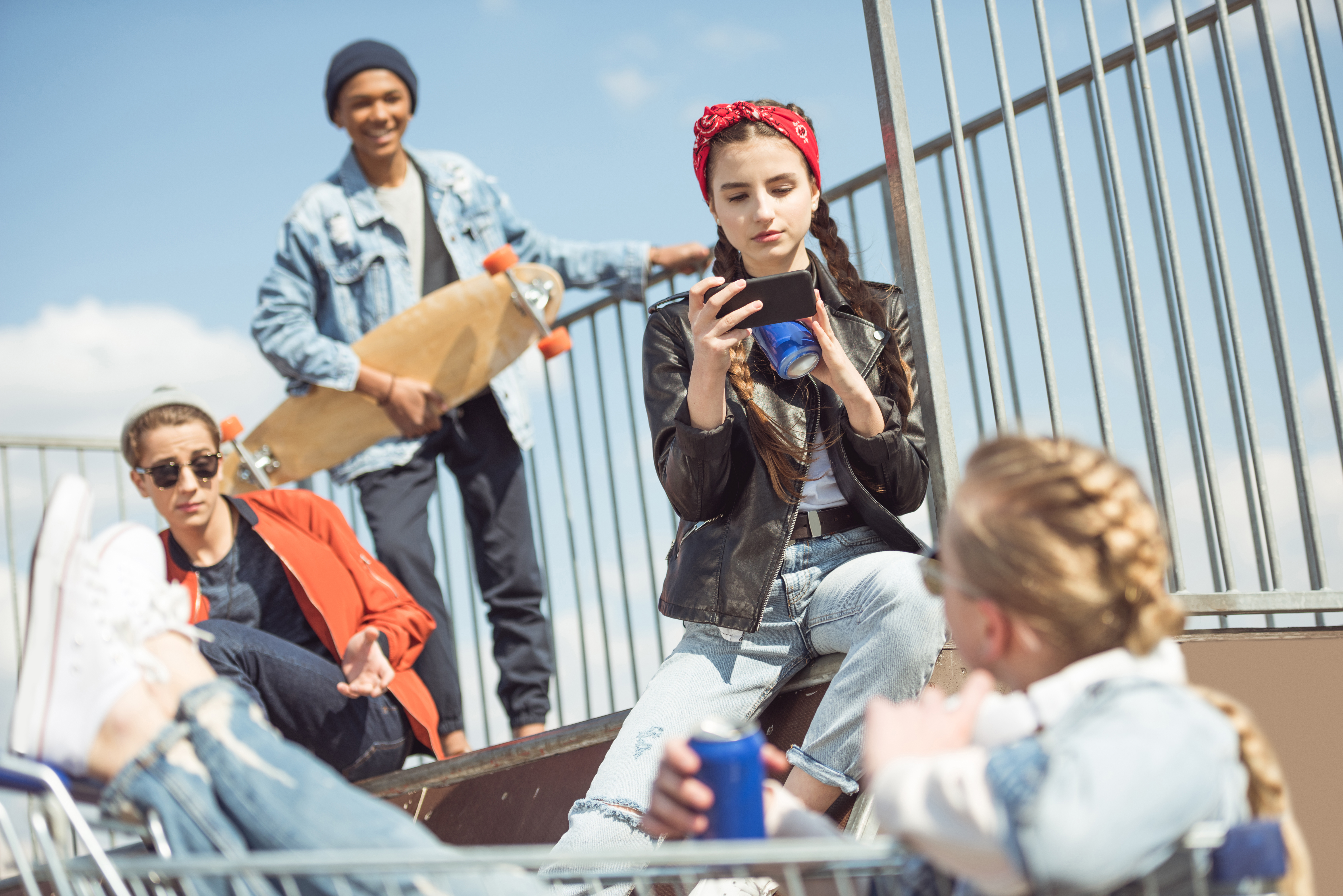 four young people wearing jackets and hats outside. one of the girls is using a smartphone to take a picture of the other girl, who is holding a can